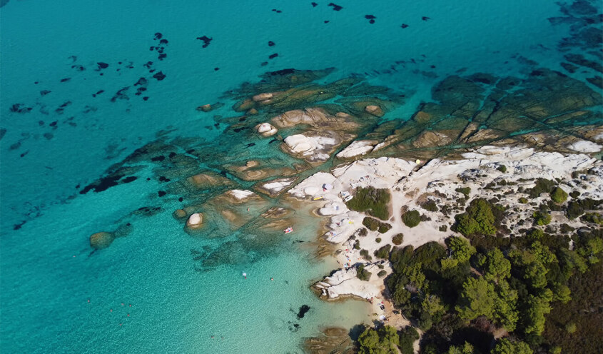 Aerial view of the crystal-clear waters and rocky shores of Orange Beach, with lush green foliage surrounding the area.