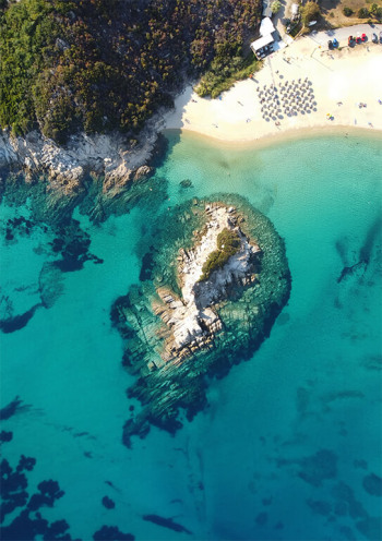 Aerial view of a tropical beach with vibrant turquoise water, a rocky outcrop, and clusters of beach umbrellas.