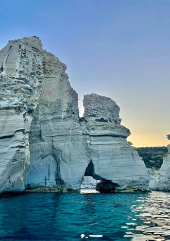 Tour boat navigating between towering white cliffs at the Kleftiko caves on Milos Island, Greece, under a serene sunset sky.