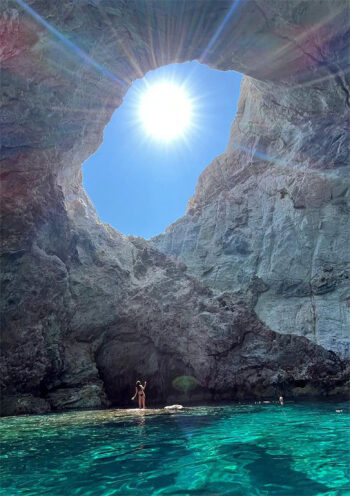 Bright sunlight shines through a gap in towering cliff walls onto a person standing on a rock in clear turquoise waters, with another person nearby, in a scenic cove.