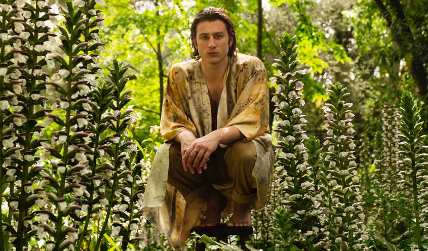 Person crouching among tall blooming flowers in a lush garden, wearing a patterned shirt and beige pants